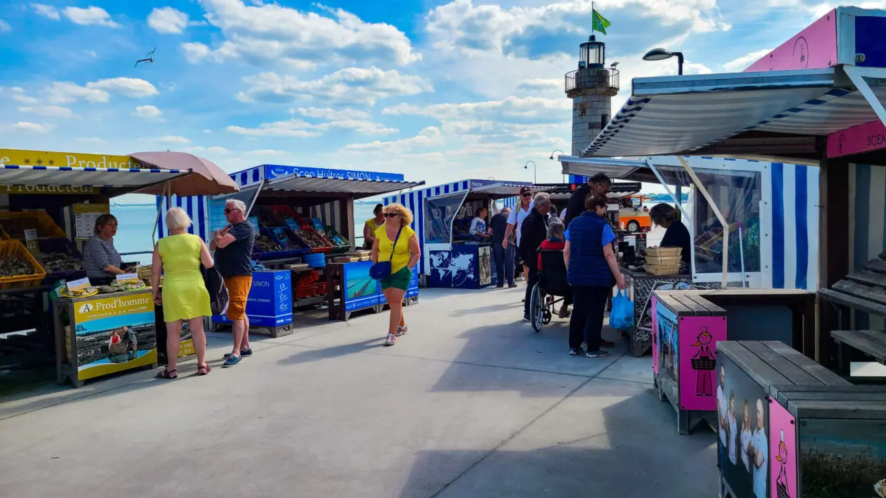 Étals de marché aux fruits de mer colorés au Marché aux Huîtres de Cancale avec phare en arrière-plan, touristes parcourant les vendeurs d'huîtres fraîches le long de la côte bretonne
